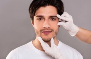 Secondary image for How Long Does a Brow Lift Last. Image of a young handsome man's chin and eyebrow being touched by gloved hands